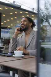 Happy businessman sitting with coffee and talking on smart phone at office park - IKF01510