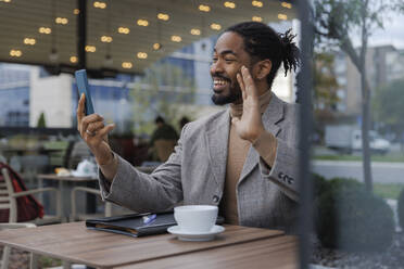 Happy businessman waving hand on video call through smart phone at cafe - IKF01512