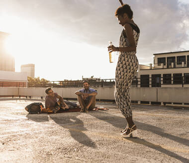 Happy woman holding beer bottle and dancing in front of friends on rooftop - UUF30767