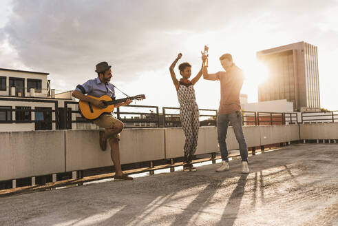 Happy man playing guitar with friends dancing on rooftop - UUF30771