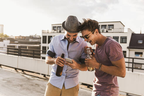 Smiling men using smart phone and holding beer bottles on rooftop - UUF30786