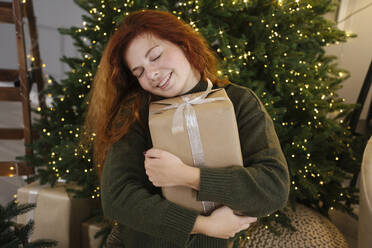 Smiling woman embracing gift box with eyes closed near Christmas tree - YBF00335