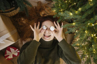 Smiling woman holding Christmas ornaments on eyes at home - YBF00336