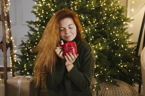 Smiling woman smelling coffee from mug near Christmas tree at home - YBF00339