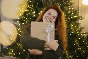 Smiling redhead woman holding gift box near Christmas tree - YBF00341
