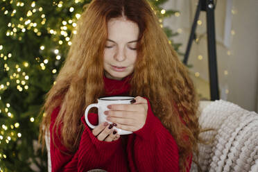 Redhead woman holding coffee mug near Christmas lights at home - YBF00343