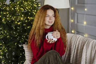 Contemplative woman sitting with coffee mug on sofa near Christmas tree - YBF00344