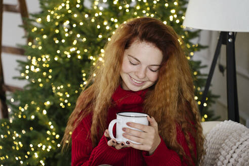 Happy redhead woman holding coffee mug in front of Christmas tree at home - YBF00346
