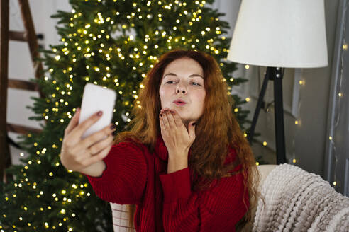 Redhead woman blowing kiss and taking selfie through smart phone at home - YBF00348