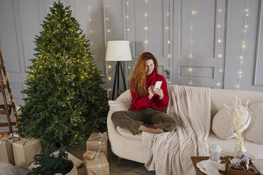 Smiling redhead woman using smart phone near Christmas tree at home - YBF00352