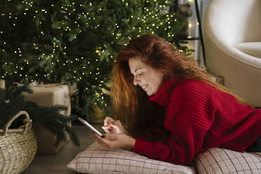 Happy redhead woman using mobile phone near Christmas tree at home - YBF00355