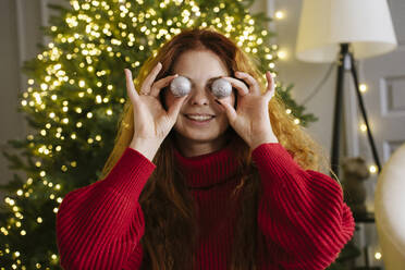 Smiling woman holding Christmas ornaments on eyes at home - YBF00356