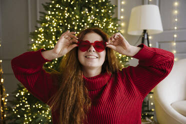 Smiling woman wearing heart shaped sunglasses near Christmas tree at home - YBF00357