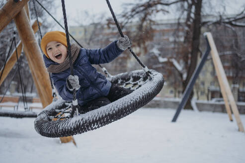 Happy boy swinging on nest swing in winter park - ANAF02536