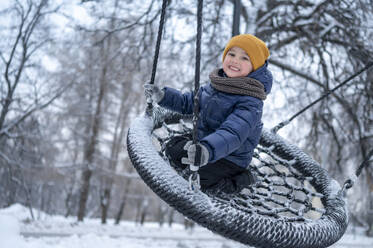 Smiling boy swinging on nest swing in winter park - ANAF02537