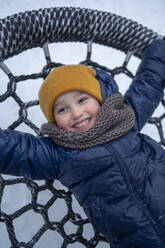 Smiling boy lying on nest swing in winter park - ANAF02539