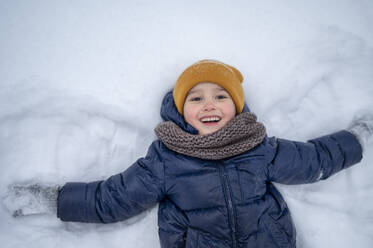 Smiling boy lying in snow in winter - ANAF02547