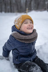 Boy sitting in snow and laughing in winter park - ANAF02548