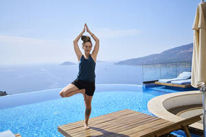 Woman doing tree pose on deck chair near swimming pool - ANNF00797