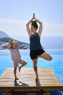 Happy mother doing tree pose with daughter near swimming pool - ANNF00800
