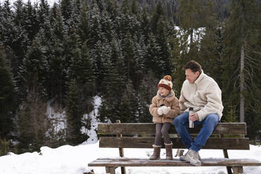 Happy daughter having tea with father on bench in winter forest - SVKF01795