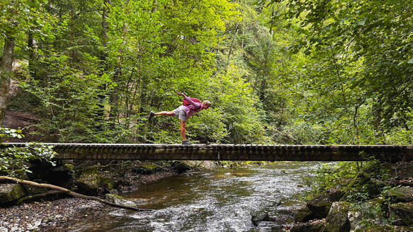 Belgien, Wallonien, Älterer Wanderer balanciert auf einem Bein auf einer Waldbrücke - GWF07960