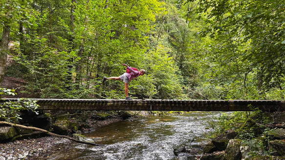 Belgium, Wallonia, Senior hiker balancing on one leg on forest bridge - GWF07960