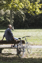 Senior man sitting on bench by bicycle in park on sunny day - UUF30788