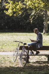 Senior man using mobile phone sitting on bench by bicycle in park - UUF30789