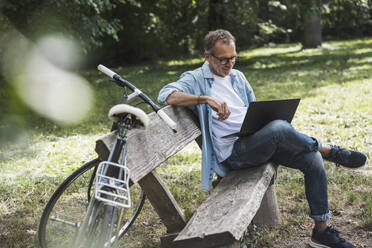 Senior man using laptop sitting by bicycle on bench in park - UUF30791