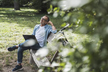 Thoughtful man with laptop sitting on bench in park - UUF30794