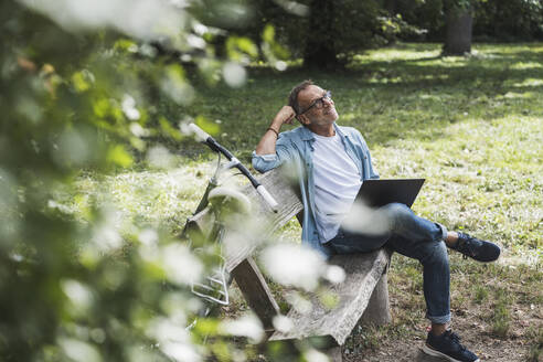 Contemplative man with laptop sitting on bench in park - UUF30795