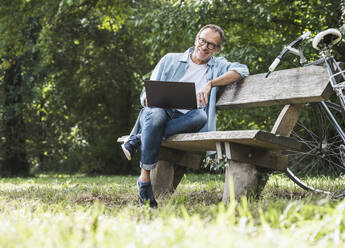 Smiling man with laptop sitting on bench in park - UUF30798