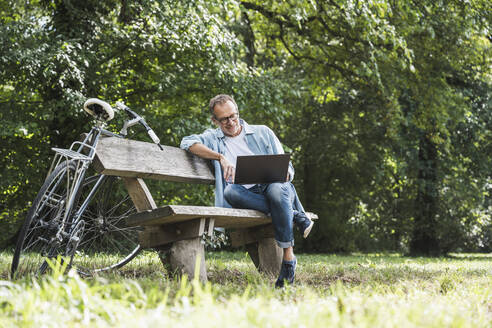 Senior man using laptop sitting on bench in park at weekend - UUF30799