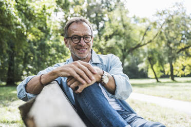 Smiling man with eyeglasses sitting on bench in park - UUF30802