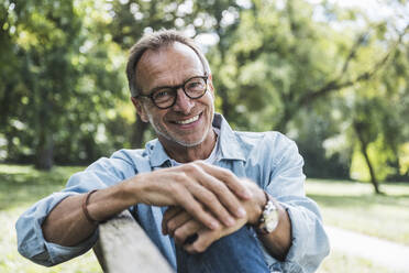 Smiling senior man with eyeglasses sitting on bench in park - UUF30803