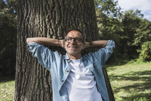 Contemplative man with hands behind head in front of tree at park - UUF30810