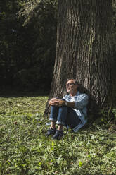 Senior man sitting in front of tree at park on sunny day - UUF30814