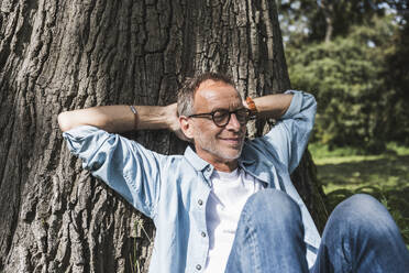 Senior man with hands behind head sitting in front of tree - UUF30817