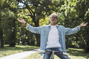 Carefree man with arms outstretched sitting on bicycle in park - UUF30821