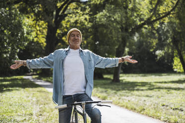 Senior man with arms outstretched sitting on bicycle in park - UUF30823