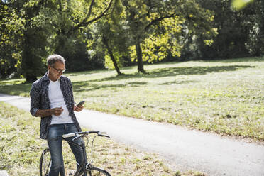 Senior man with bicycle using mobile phone in park - UUF30824