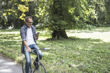 Senior man riding bicycle and using smart phone in park - UUF30825