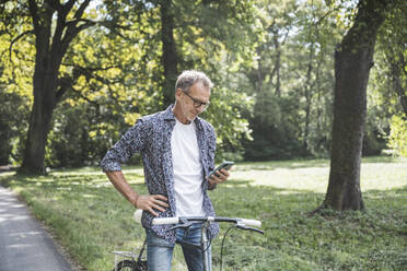 Senior man with bicycle using smart phone in park - UUF30826