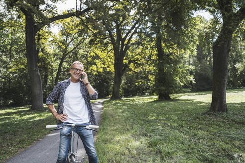 Senior man with bicycle talking on smart phone in park - UUF30827