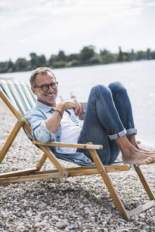 Smiling senior man sitting on deck chair at beach - UUF30828