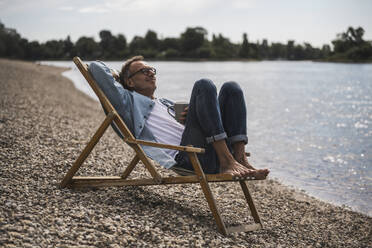 Senior man with coffee cup relaxing on deck chair by sea - UUF30830