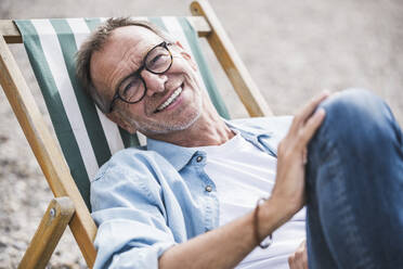 Happy senior man sitting on deck chair at beach - UUF30833