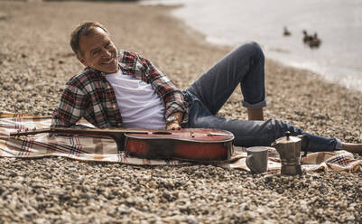 Smiling man with guitar sitting on blanket at beach - UUF30840