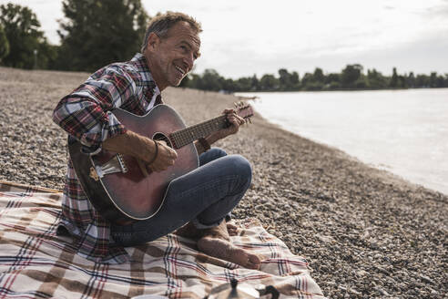 Smiling senior man playing guitar and sitting on blanket at beach - UUF30841